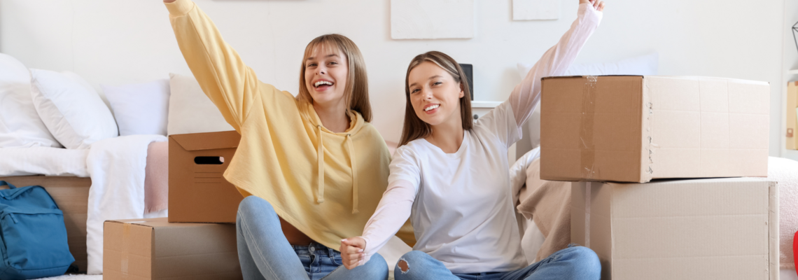 Abbildung zwei jugendliche Mädchen sitzen am Boden, neben ihnen stehen volle Umzugkartons.