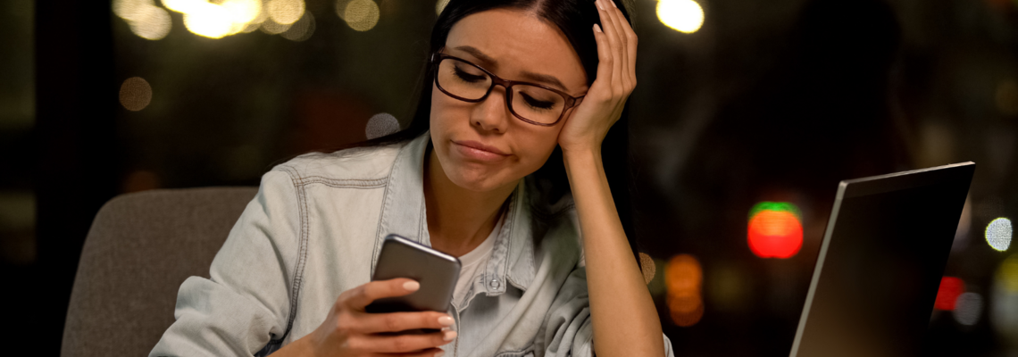 Abbildung eine Jugendliche sitzt an ihrem Schreibtisch und hat ihr Handy in der Hand, während sie sich auf ihren aufgelehnten Laptop abstützt.